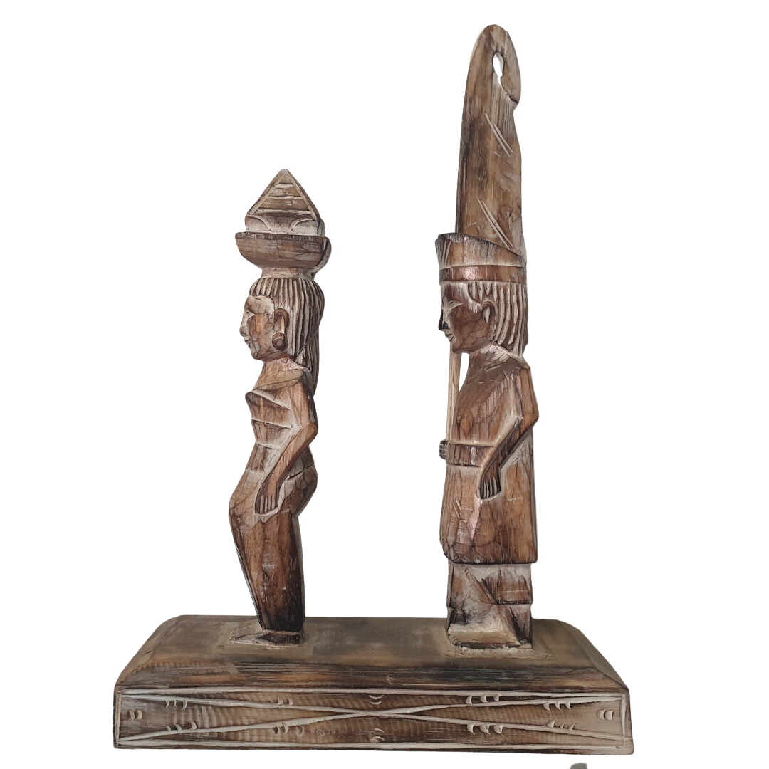 A wooden statue depicting two Balinese figures in traditional ceremony attire, standing on a platform.