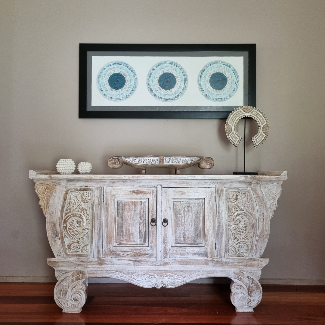 Balinese teak entryway cabinet styled with carved timber decorative objects and framed coastal wall art above, warm interior setting