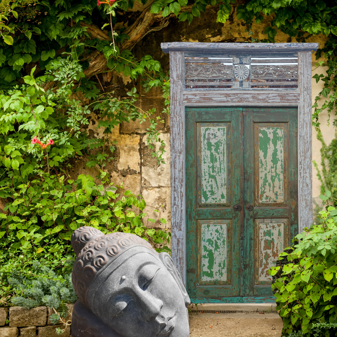 Decorative wooden door with greenish patina in a garden setting with a Buddha statue and foliage.