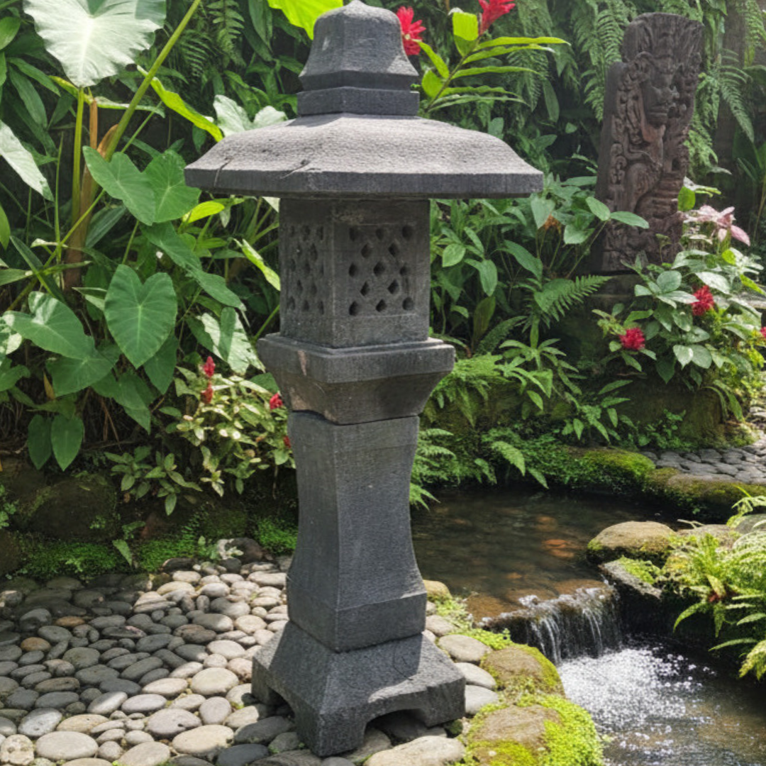 Lava stone garden lantern with round perforated light chamber and tapered column positioned on a river pebble path beside a cascading stream in a lush tropical Balinese garden with ferns, elephant ears and red heliconia