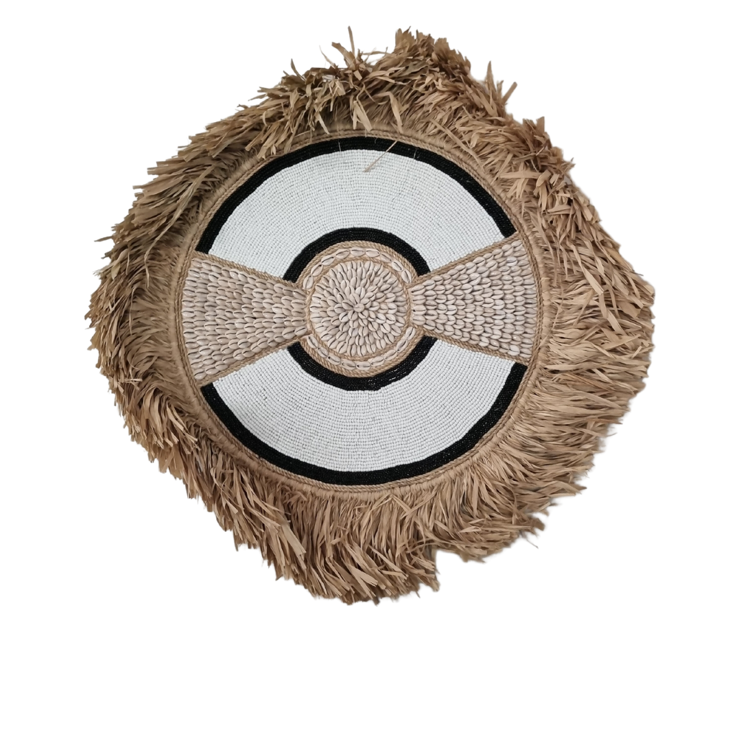Round shell-and-bead piece with black circular bands, shell panels at either side and tan raffia fringe against a white background