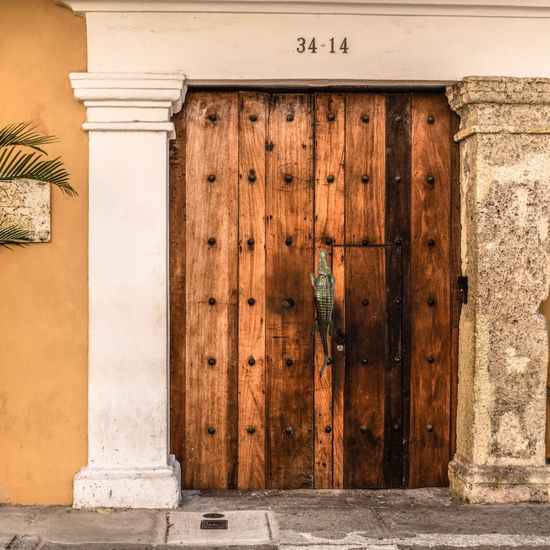 Solid brass crocodile door handle mounted on rustic studded double timber doors – white colonial archway with warm ochre rendered walls – decorative brass hardware Australia