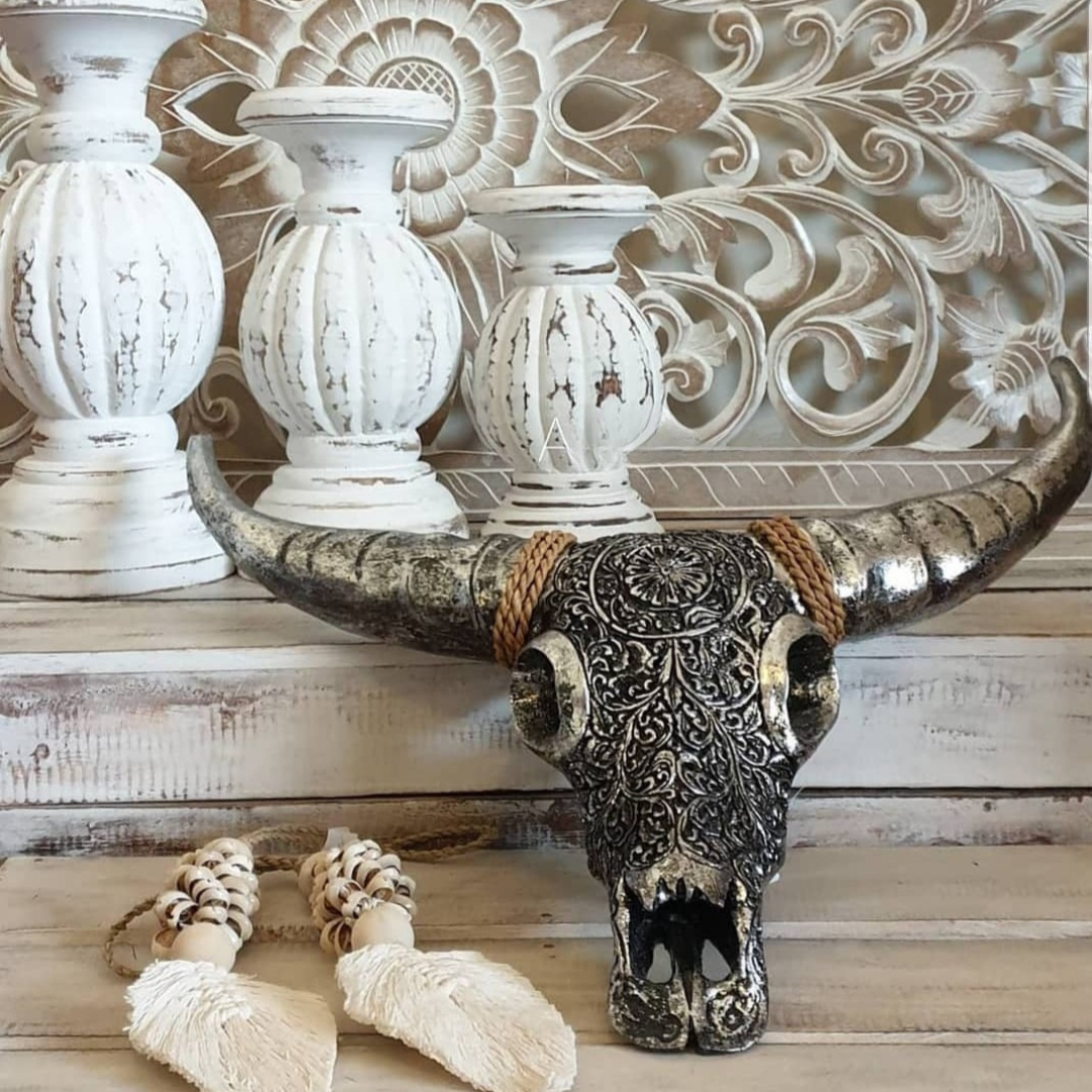 Metallic silver bull skull wall art with intricate mandala engraving styled on a whitewashed timber tabletop with distressed white candle holders, macramé beads and carved floral wall panel — boho rustic home décor by Azure Decor