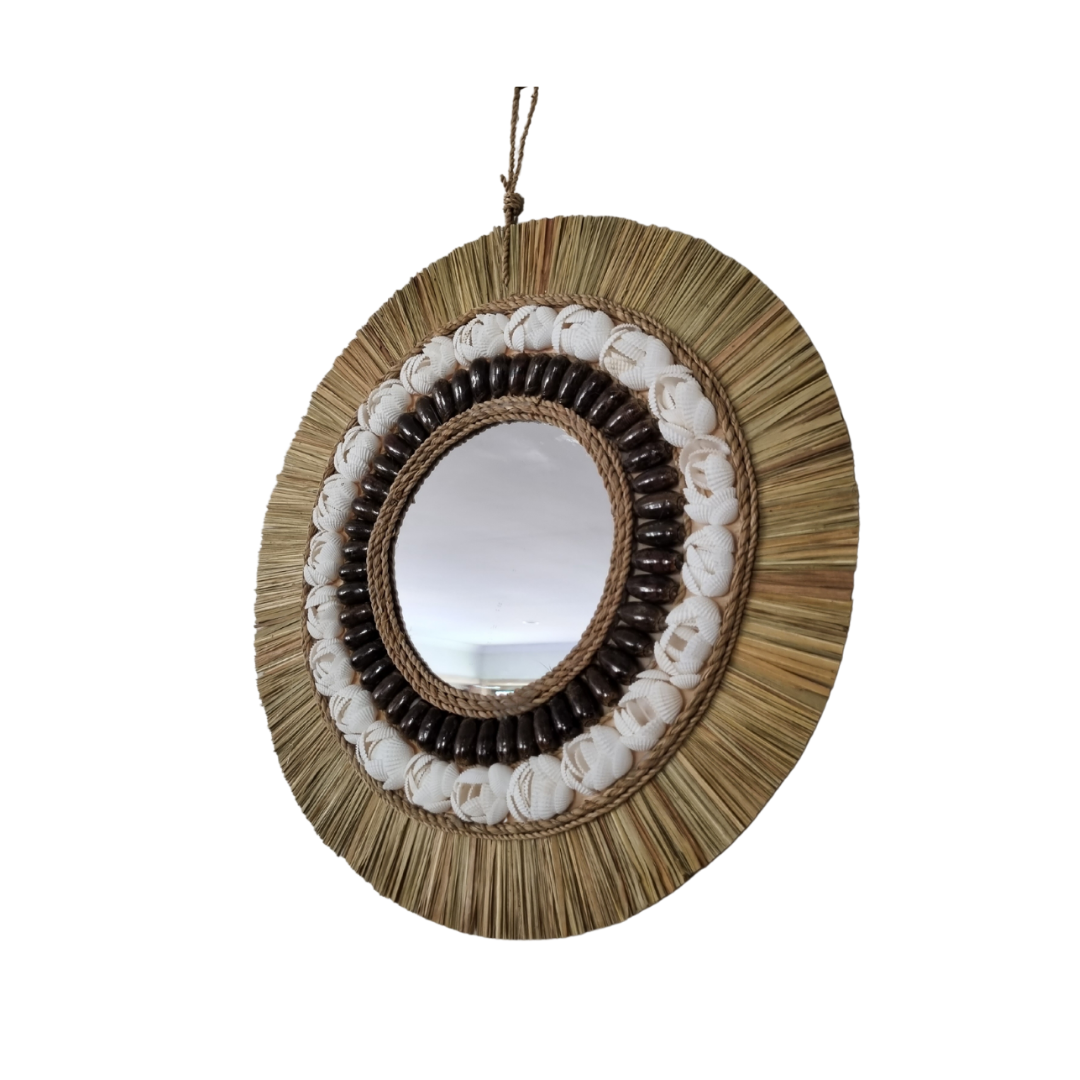 Round coastal boho wall mirror angle shot with natural seagrass raffia fringe, white shells and dark shell, styled above a slim console table in a bright entryway with monstera plant and coastal decor — handcrafted coastal boho home decor by Azure Decor