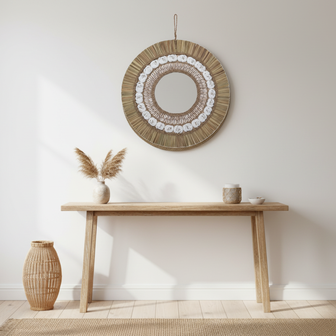 Round natural-fibre frame with pale decorative details above a timber console, styled with pampas stems, neutral ceramics and a woven floor vase