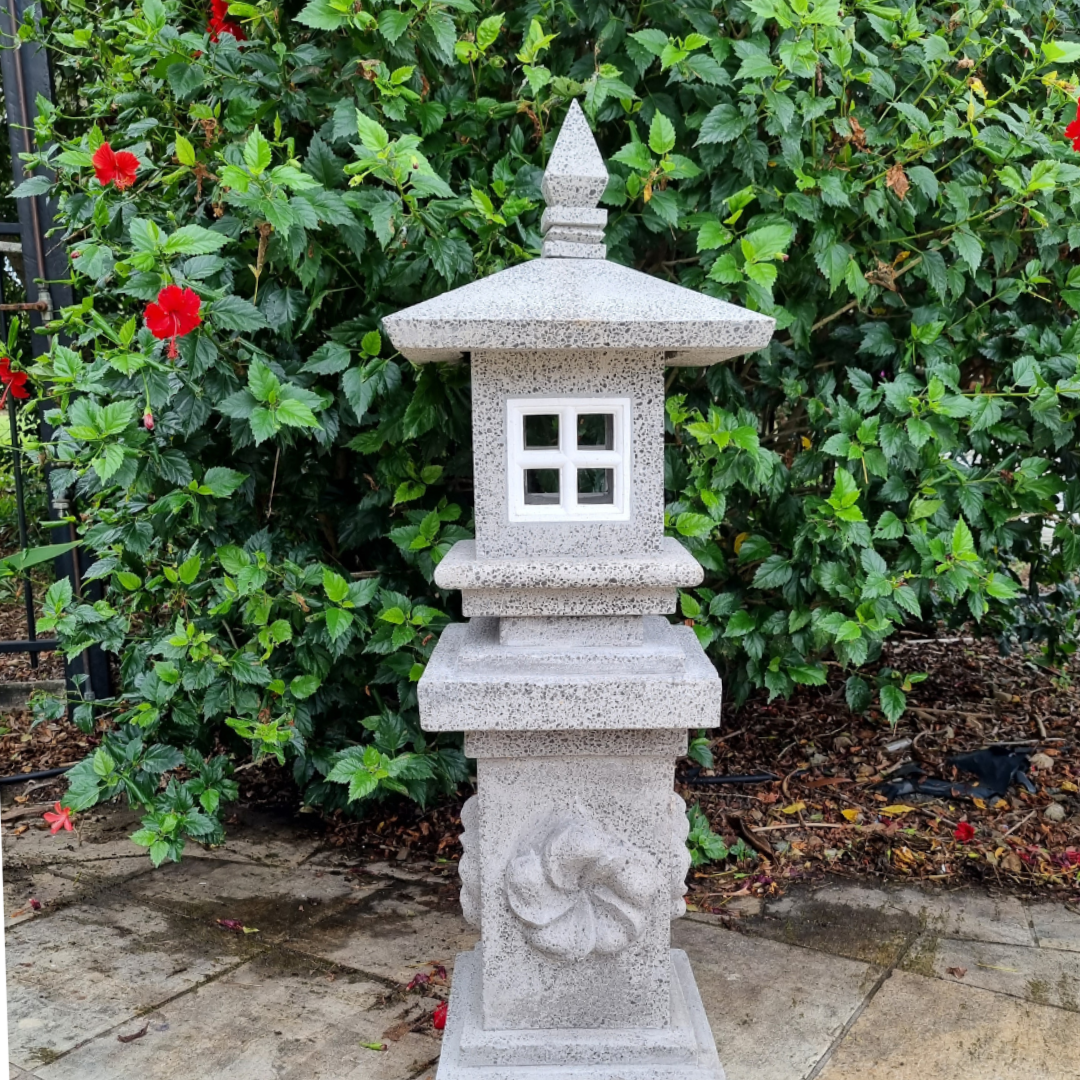 Tall concrete pagoda lantern in light grey with carved spiral detail on the column, square light opening and pointed finial, styled against a lush green hibiscus hedge