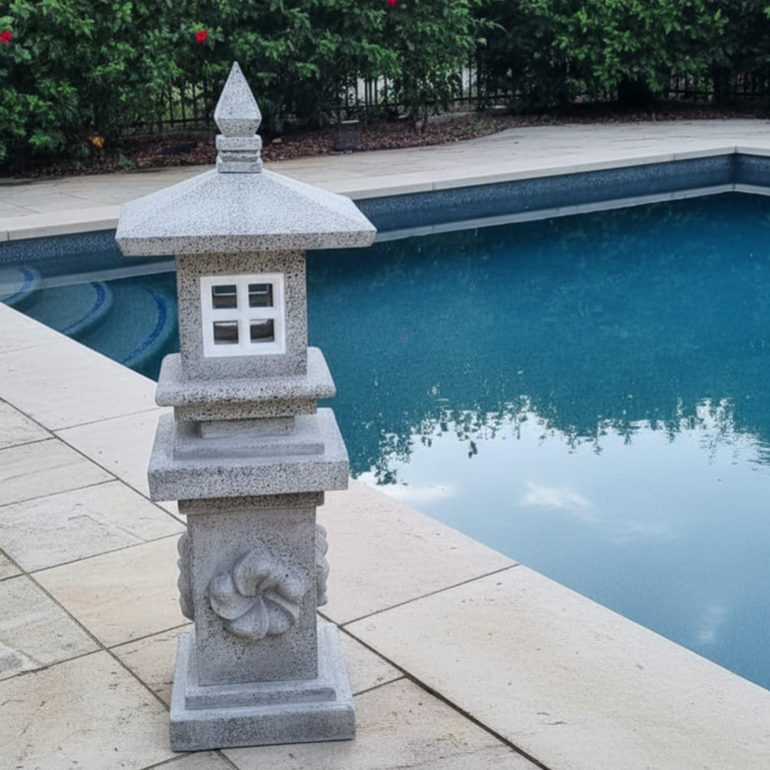 Concrete pagoda lantern with carved spiral column and square light opening positioned on travertine pool coping beside a blue swimming pool with lush green hedging