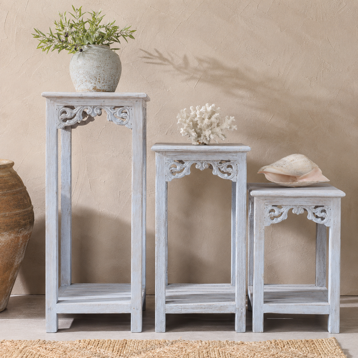 Three distressed white wash wooden nesting side tables of graduated sizes, styled with a ceramic pot, coral branch, and seashell against a textured beige wall.