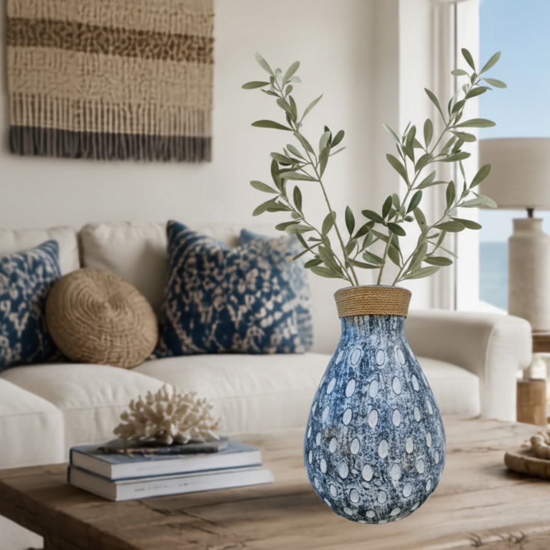 Teardrop-shaped pot with dark blue-grey distressed finish and raised oval dots, woven jute collar at the opening, filled with olive branches, styled on a driftwood coffee table in a bright coastal living room with ocean views