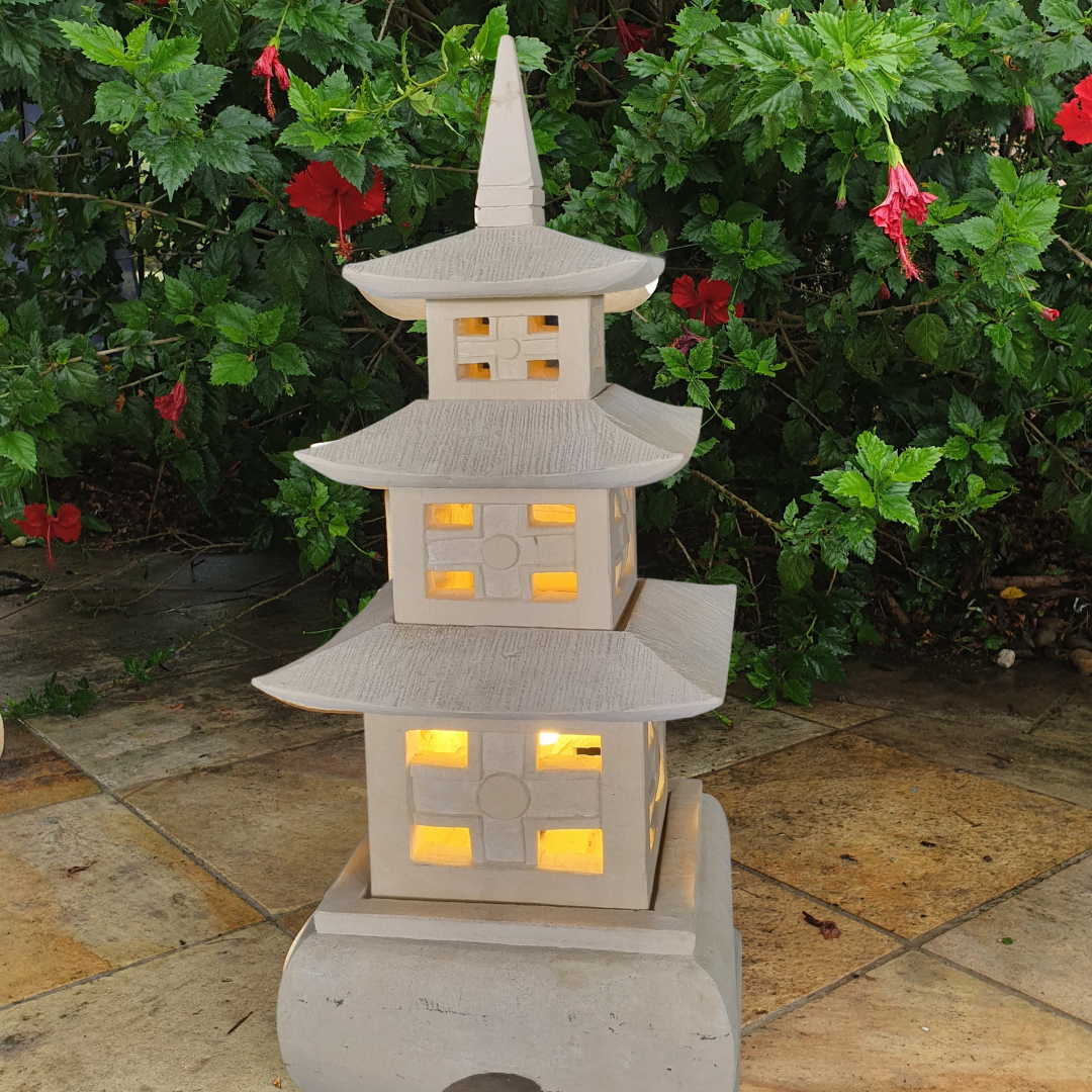 Stone pagoda lantern with amber-lit carved windows on a sandstone patio, red hibiscus and green foliage in the background