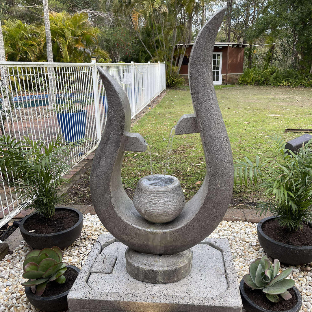 Decorative stone sculpture in a garden setting with plants and a fence in the background.