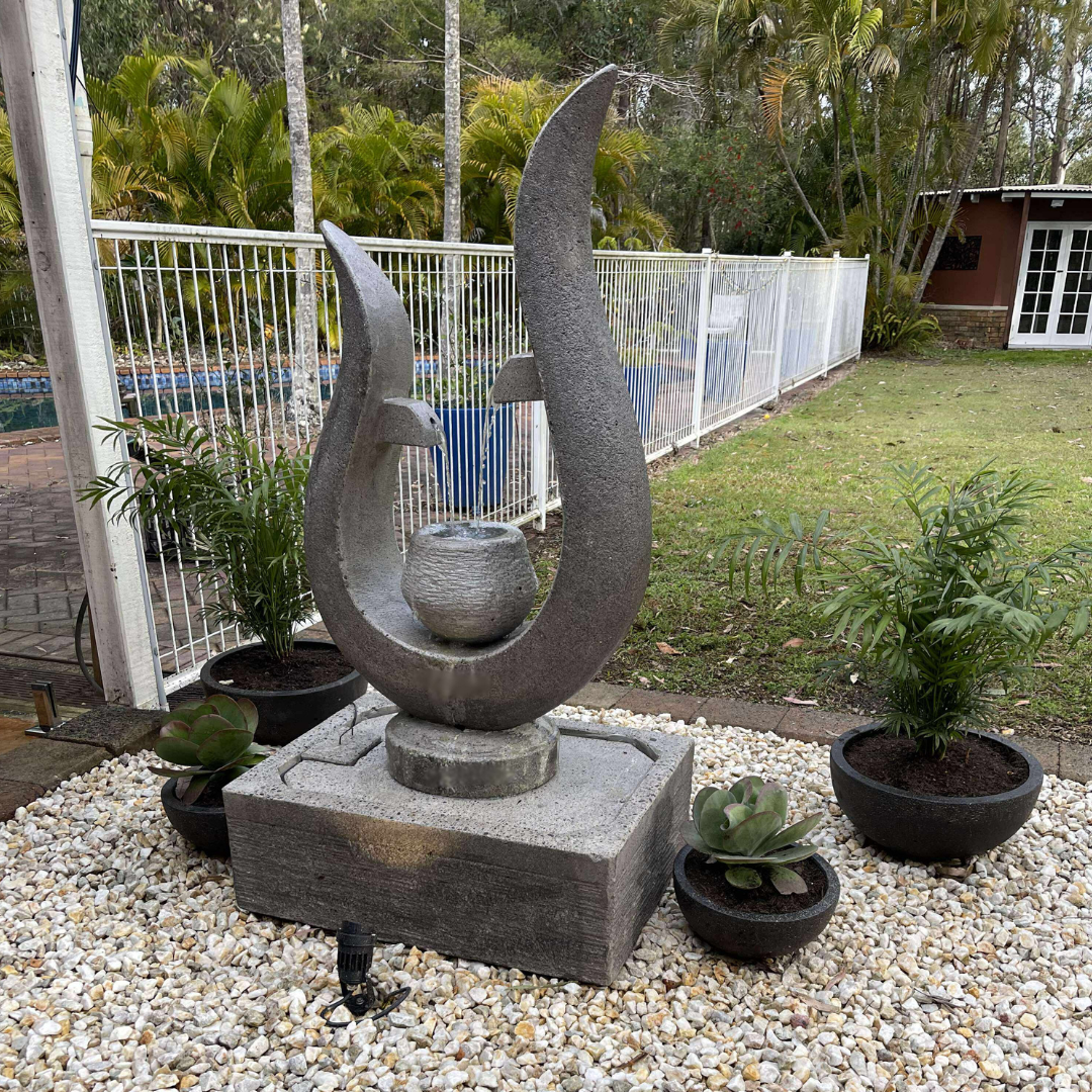 Decorative stone sculpture in a garden setting with plants and a pool in the background.