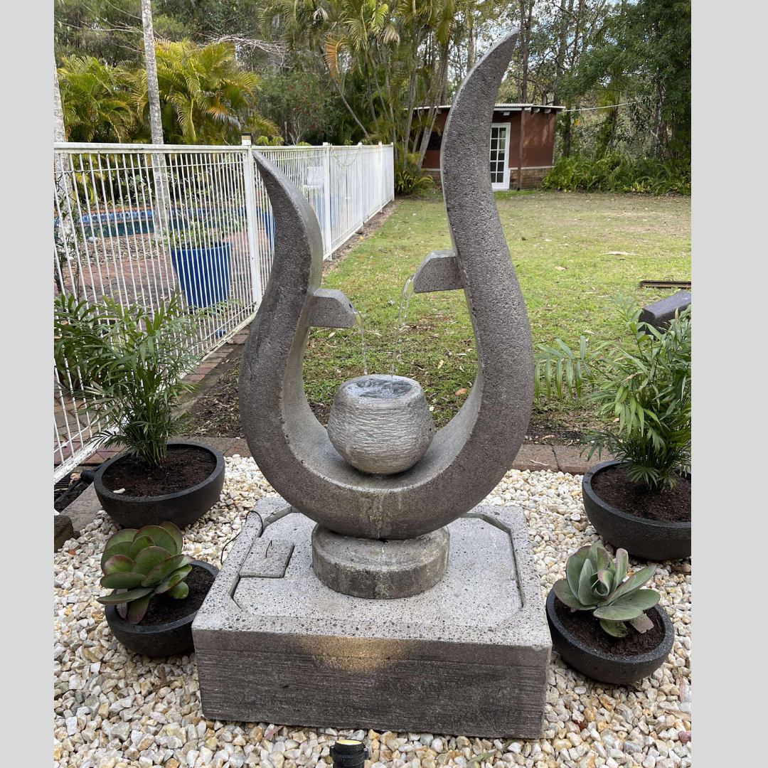 Stone sculpture of two curved stones with a small bowl in the center, set against a garden backdrop.