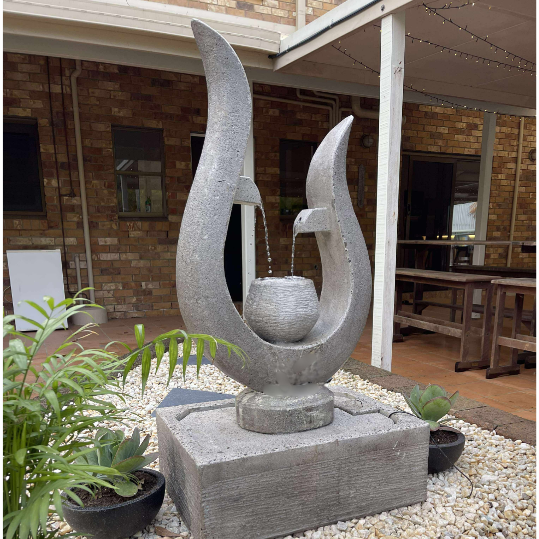 Decorative stone water fountain in a garden setting with plants and a wooden building in the background.