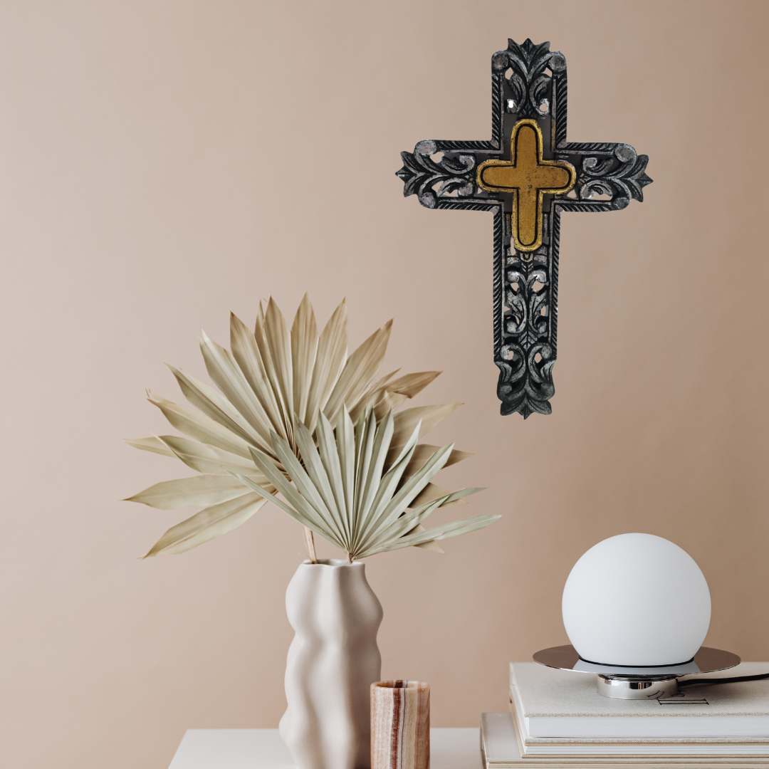 Decorative cross on a wall above a table with a vase, plant, and lamp.