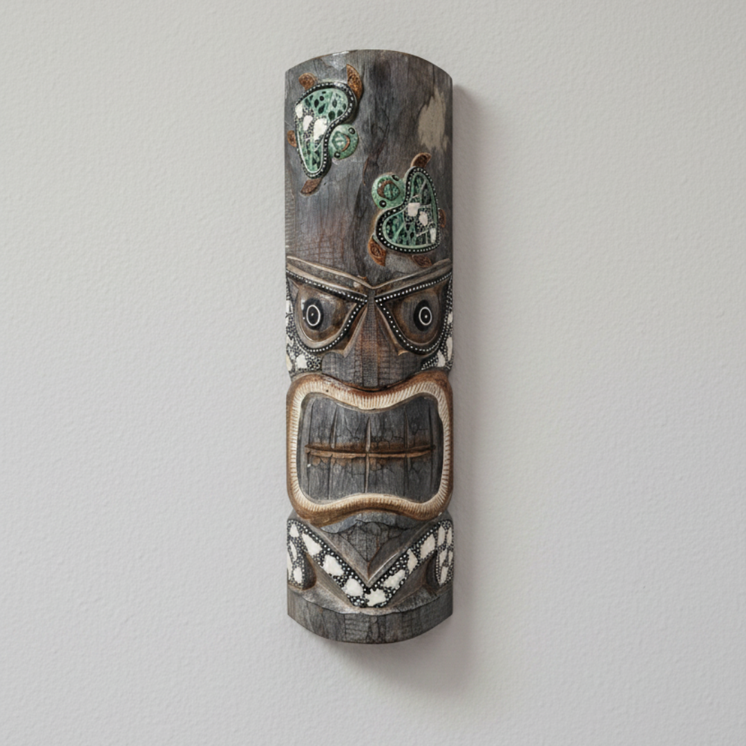 Hand-carved wooden tiki mask with sea turtle motif mounted flush on a neutral wall, showing dot-work eye detailing, open rectangular mouth, and shell grit chevron accents in weathered dark grey and brown tones.