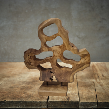 Teak Wood Table Top Sculpture displayed on a rustic timber surface against a grey concrete wall, showing natural teak grain and organic abstract form