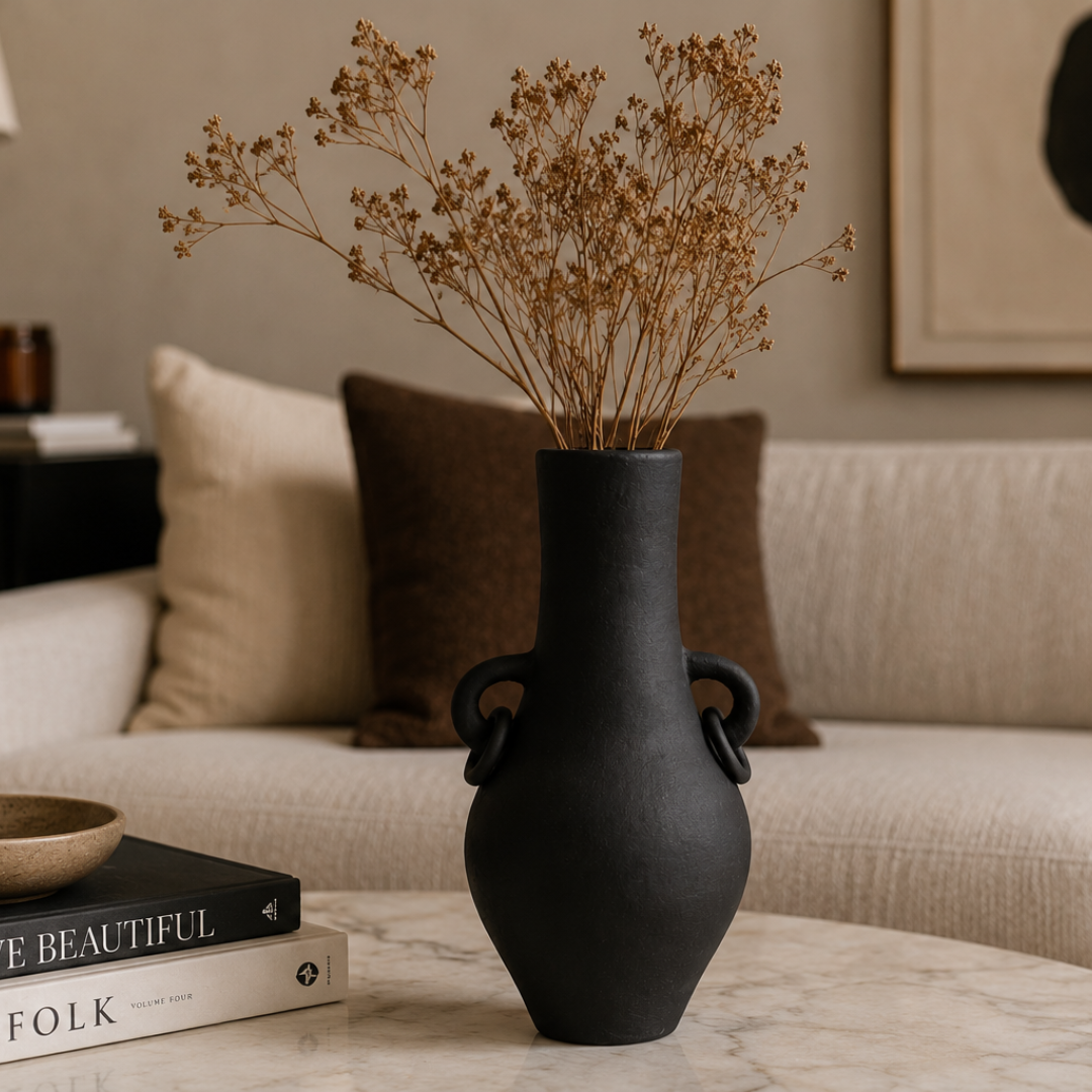 Handcrafted terracotta vase with ring handles in matte black styled with dried branches on a marble coffee table beside stacked books in a warm neutral living room