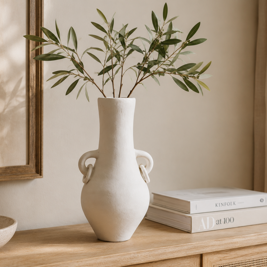 Matte white terracotta vase with looped ring handles styled with olive branches on a timber sideboard beside stacked books and a stone bowl in a warm coastal interior