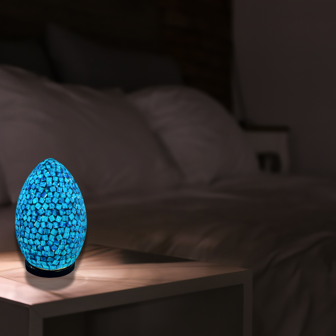 Art Deco oval mosaic table lamp in vivid blue, glowing on a timber bedside table against a dark bedroom backdrop