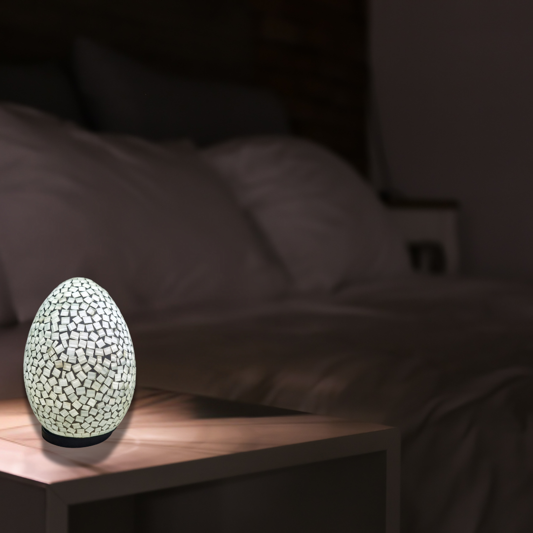 Art Deco oval mosaic table lamp in white, illuminated on a timber bedside table in a softly lit bedroom
