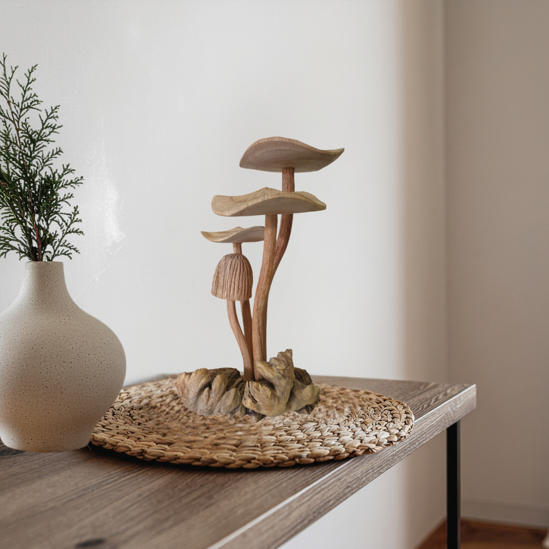 Wooden mushroom decor on a woven mat with a vase containing greenery on a wooden surface.