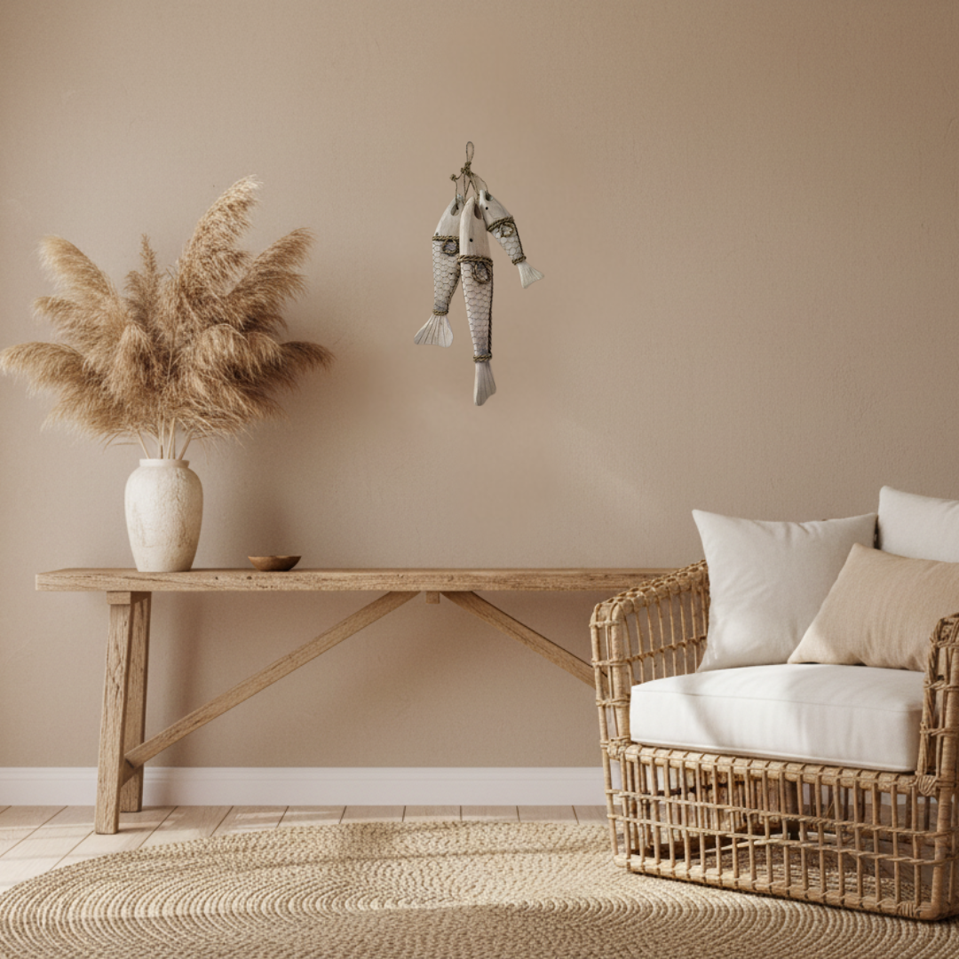 A styled interior scene with a rattan armchair, white and beige cushions, a light wood console table, a vase of pampas grass, and a hanging fish decoration against a warm beige wall.