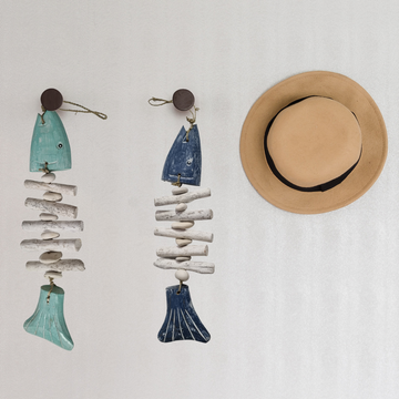 Two decorative fish ornaments made from driftwood and ceramic on a white background, with a straw hat beside them.