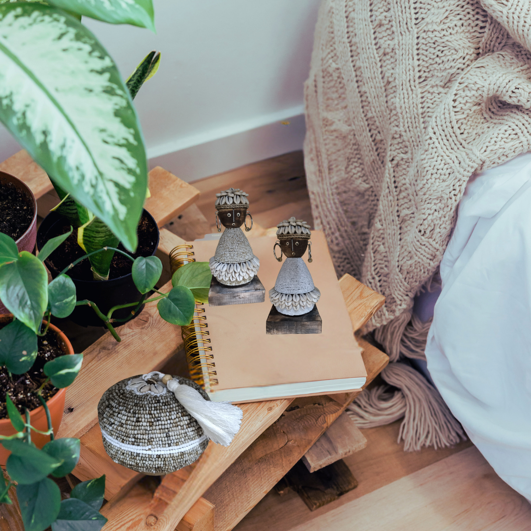 WOODEN DOLL SET IN A BOHO BEDROOM STYLE OF HOME SITTING ON A BOOK WITH GREEN PLANTS AND BEADED BASKET 