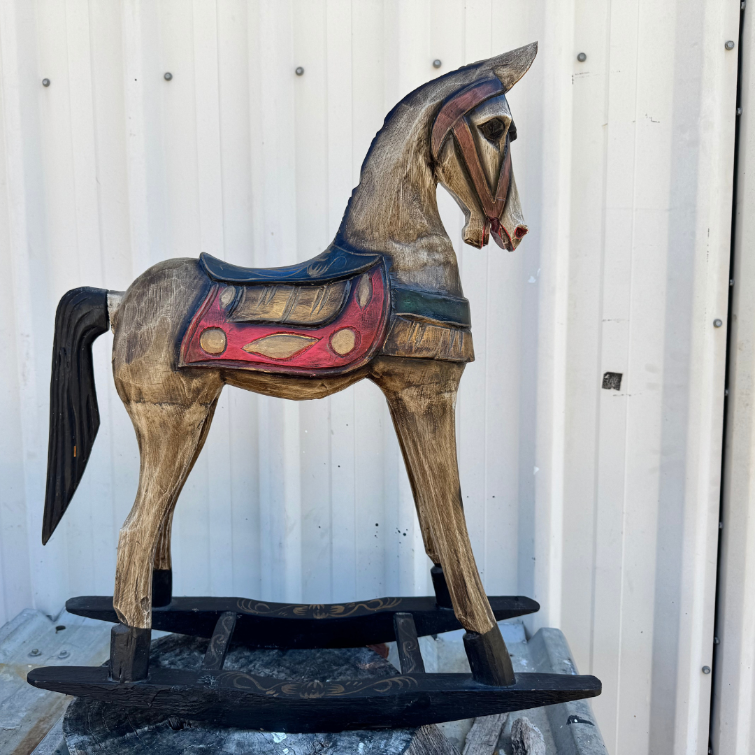 Rustic timber horse sculpture with weathered tan-brown finish, red and gold saddle detailing, dark carved mane, and black rocker base, against a white corrugated metal wall.