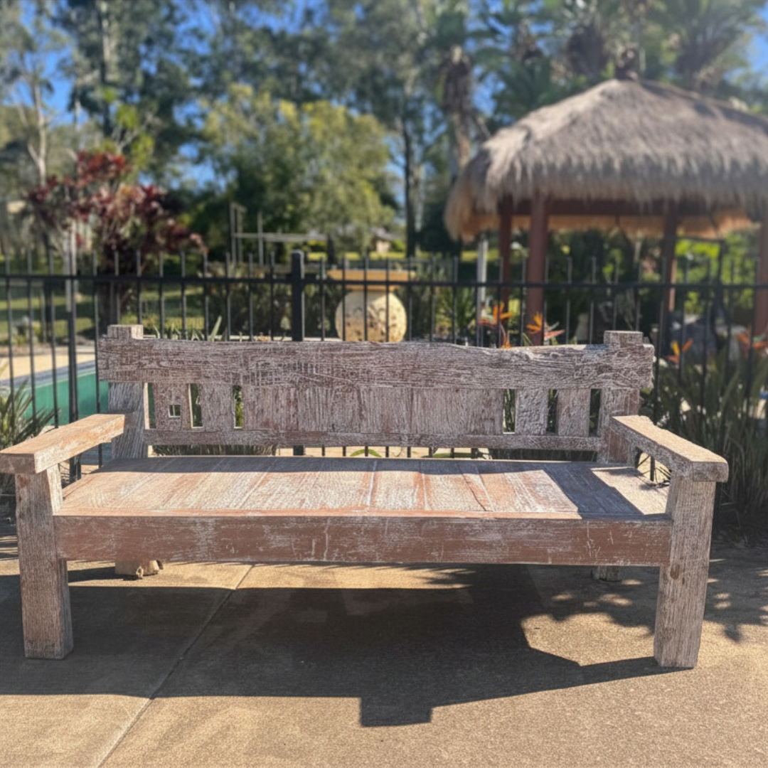 Weathered timber bench seat on a sunny poolside patio, with a thatched Balinese hut, tropical plants and iron fence in the background