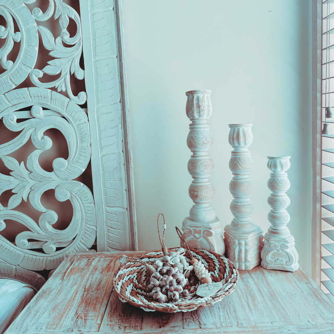 Small seagrass spiral piece styled flat as a tabletop bowl/tray with shells and beads inside, next to white carved candlesticks and a Balinese carved panel — beautiful coastal Hamptons vignette.