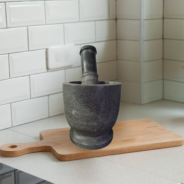 Stone mortar and pestle set surrounded by fresh herbs and whole spices including star anise, chilli, cardamom, and bay leaves on a timber board