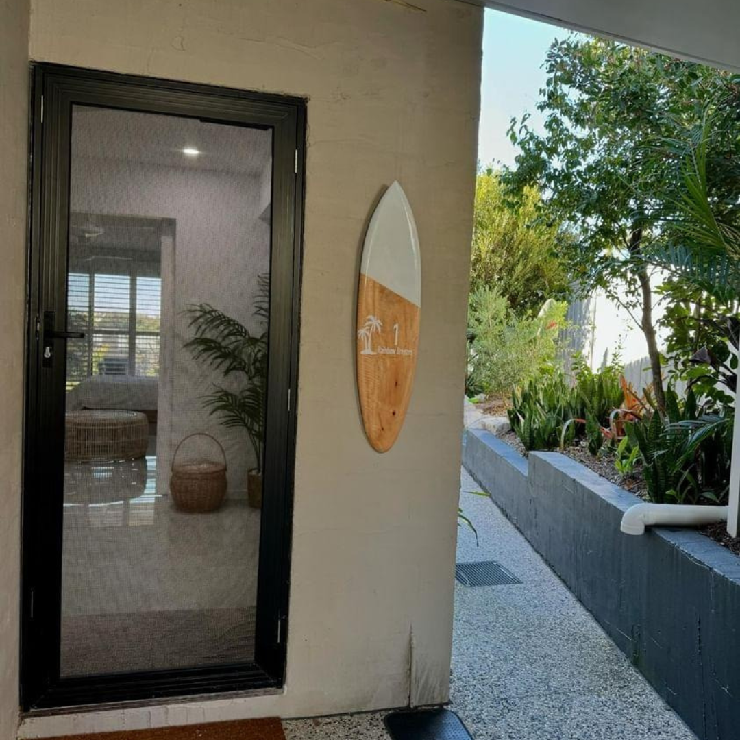 set undercover. Sign mounted beside a front door on an exterior wall, white painted top half with palm tree engraving and house number, tropical garden in background