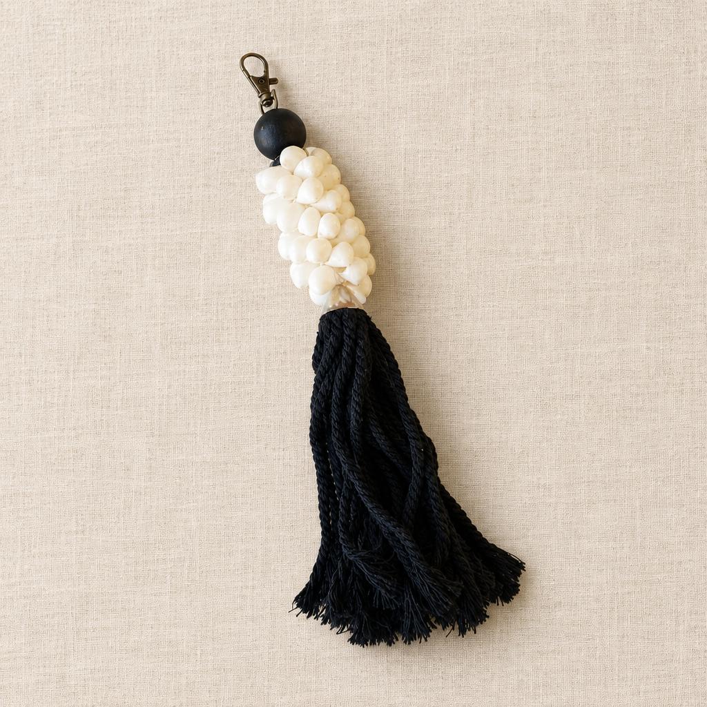 A handmade keychain or bag charm featuring a black spherical bead, a cluster of small white beads shaped like a corn cob, and a long black braided rope tassel, displayed on a textured beige linen background.