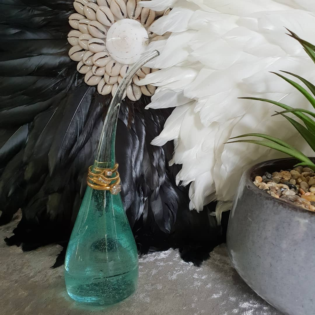 Small aqua teal glass sculpture with gold rings on the stem styled in front of a black and white Juju feather wall art with cowrie shells and an air plant