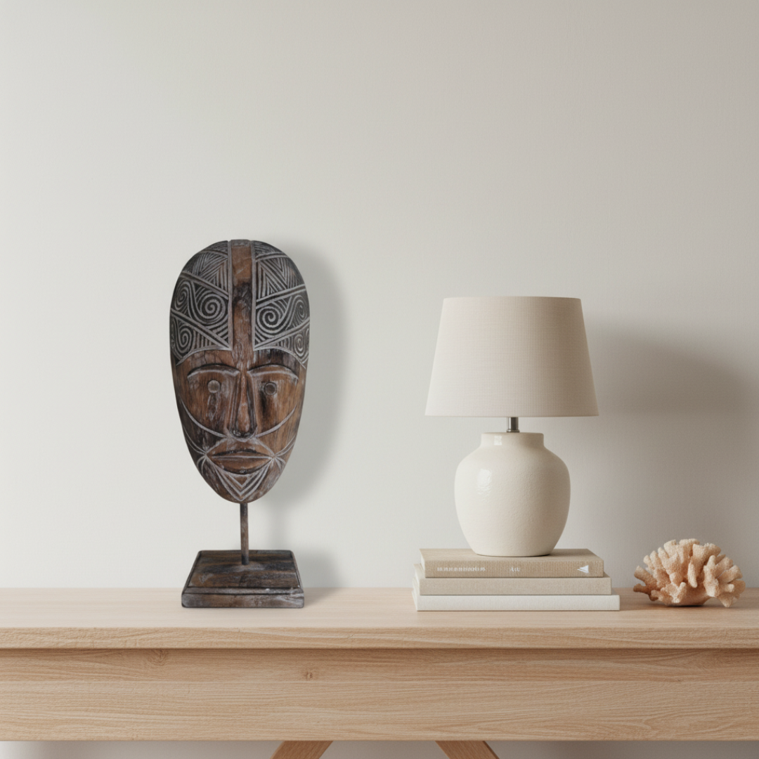 A carved wooden mask on a metal stand displayed on a light wood console table next to a cream table lamp on stacked books and a coral shell, against a neutral off-white wall.
