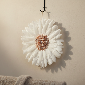 Handcrafted round white feather wall hanging with a blush pink spiral shell centrepiece hung on a hook against a warm cream wall with a grey linen throw in the foreground