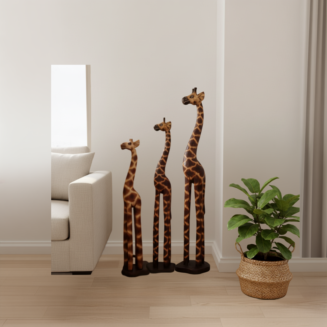 Set of three carved timber giraffe figurines in small, medium and large sizes grouped together in a coastal lounge room corner beside a large indoor floor plant in a woven basket pot, with a linen sofa and warm natural light