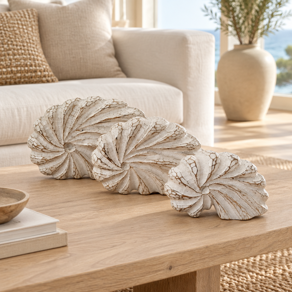 Three distressed white seashell-shaped decorative sculptures arranged on a light wood coffee table in a bright, modern living room.