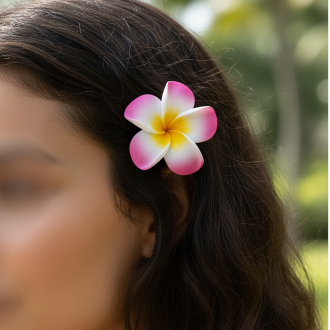 Pink and white frangipani flower hair clip worn in dark hair, blurred green palm trees in the background