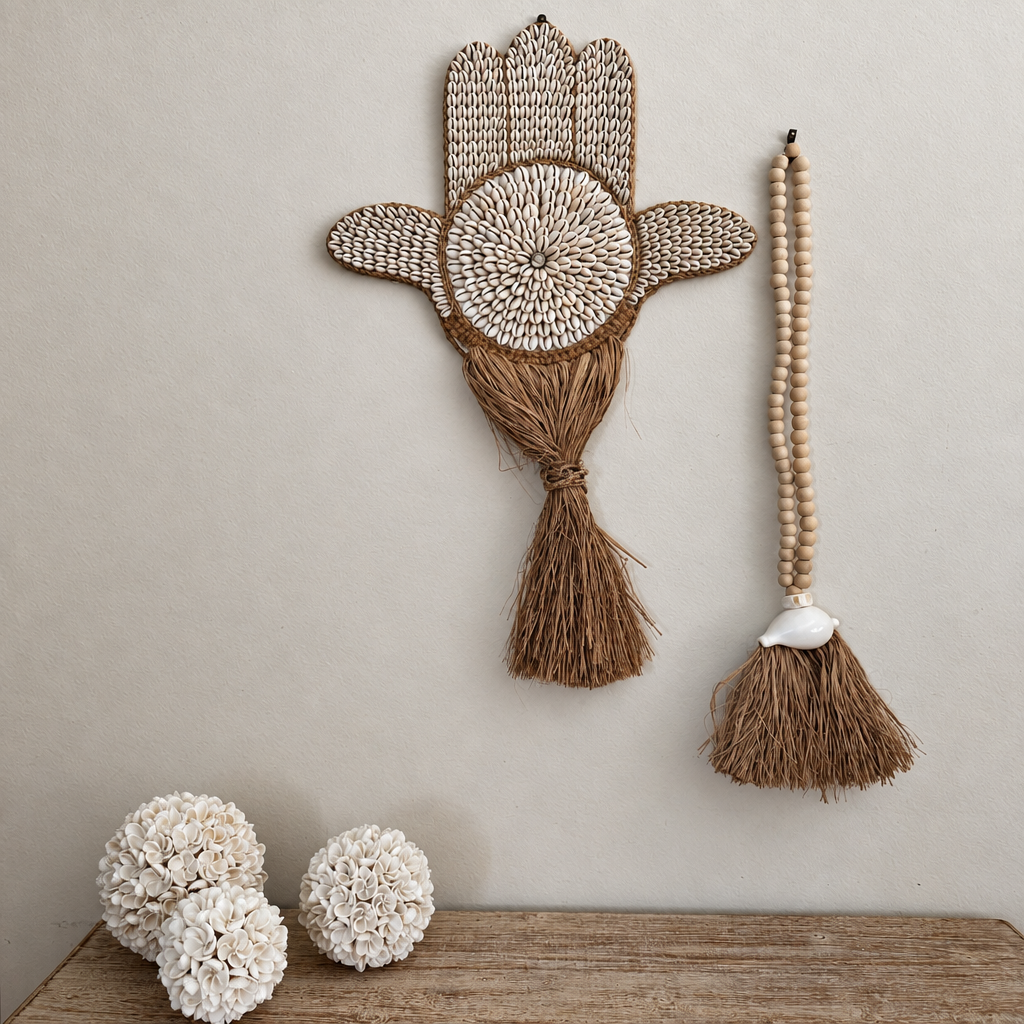 Cowrie shell hand-shaped wall hanging beside a long shell tassel decoration on a pale wall above a timber console
