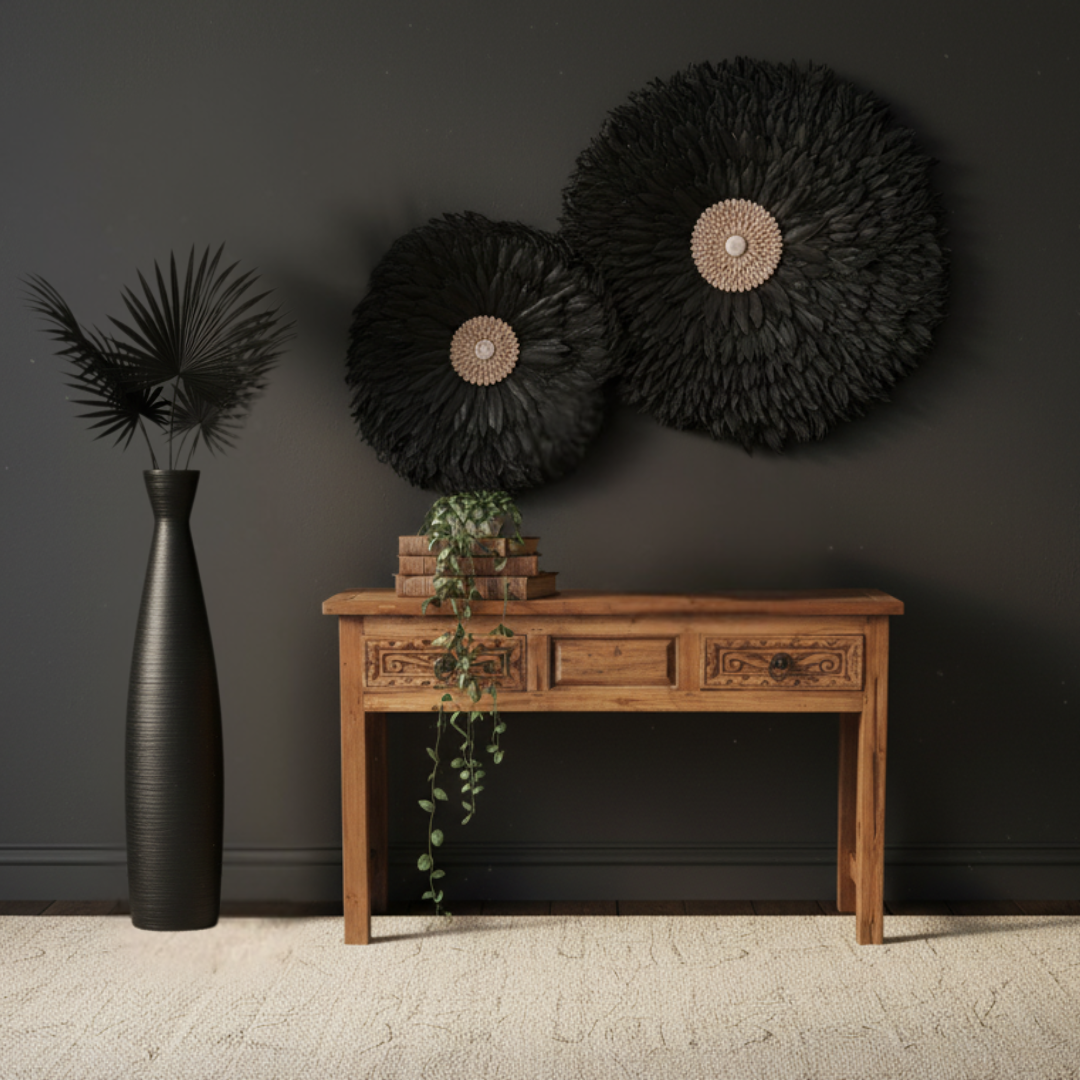 Two large round black feather wall discs with shell centres styled above a carved timber console table with trailing greenery and a tall black vase against a dark charcoal wall