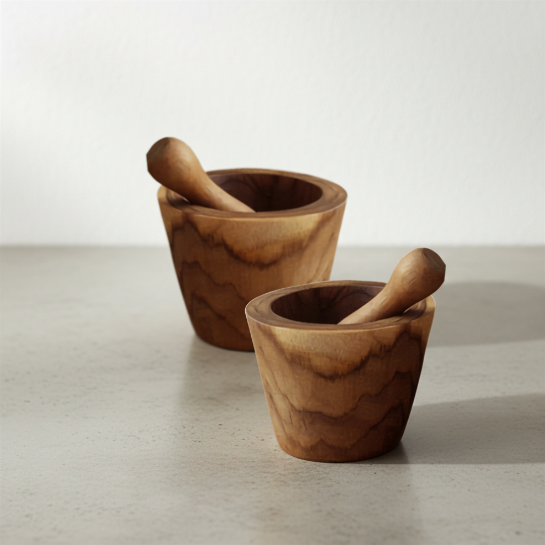 Two handcrafted teak wood mortar and pestle sets in cylindrical style, styled on a grey concrete surface against a soft white wall, showing rich swirling wood grain