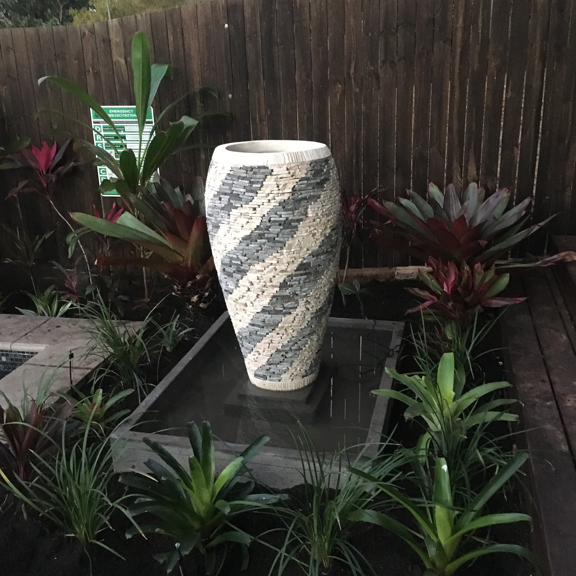 Marble and pebble Balinese style garden pot set on a dark reflective platform surrounded by tropical bromeliads and lush garden plants, with a timber fence backdrop