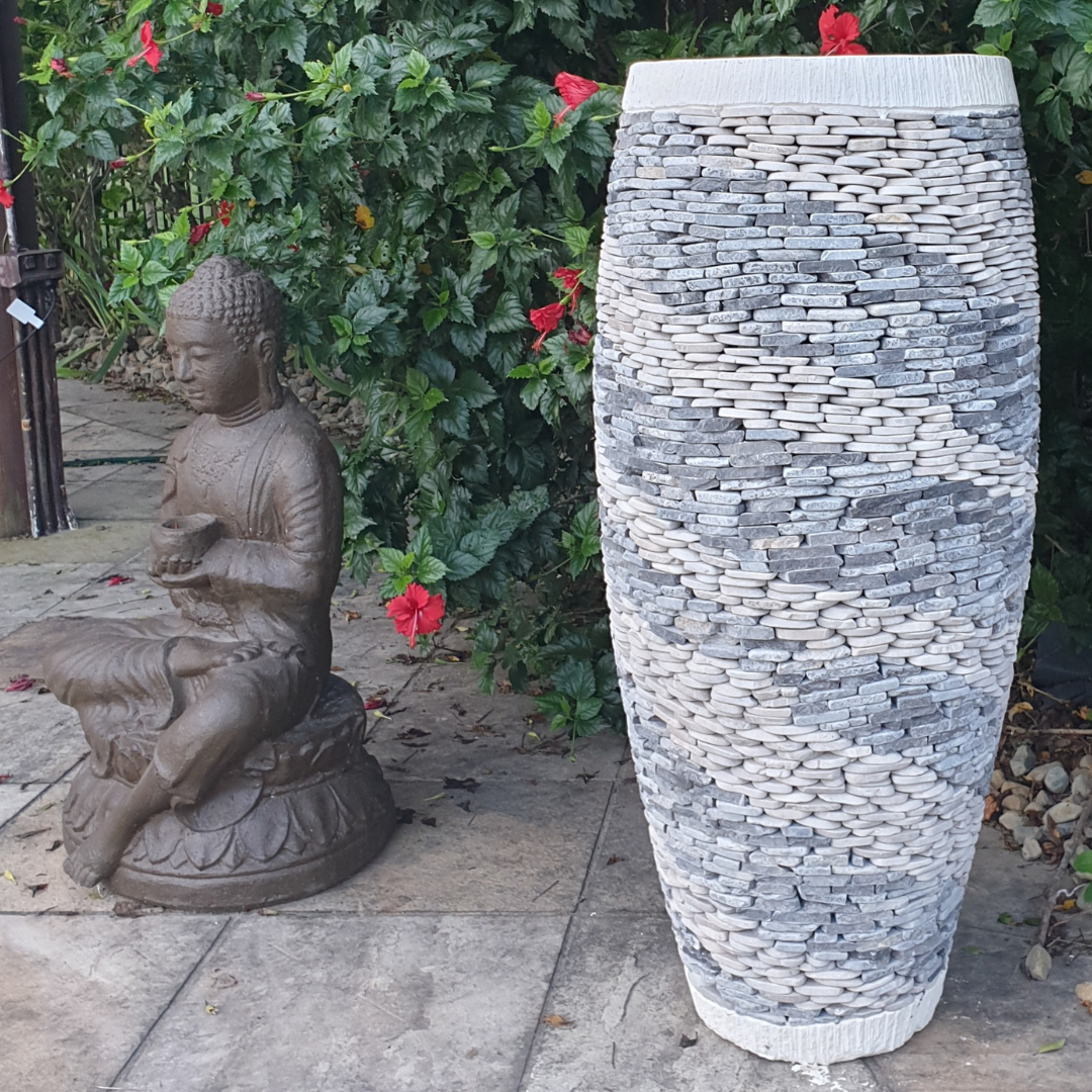 Marble and pebble Balinese style garden pot displayed on a stone patio beside a seated Buddha statue, with lush green foliage and red hibiscus flowers in the background